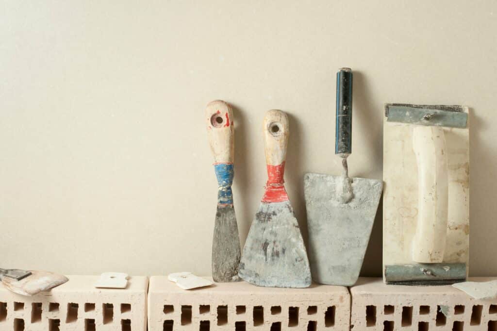 Des outils pour appliquer l'enduit alignés sur un mur de couleur beige et au dessus de briques.