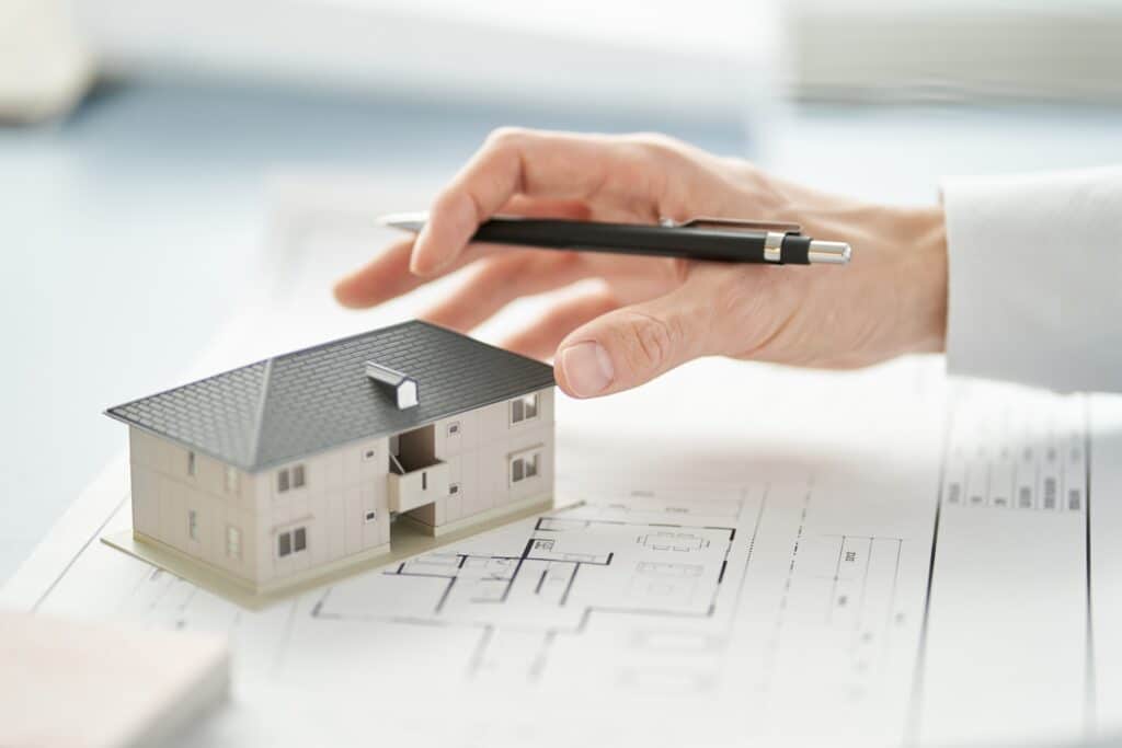 Les mains d'un homme tenant un stylo et touchant la maquette d'une maison posée sur un plan.