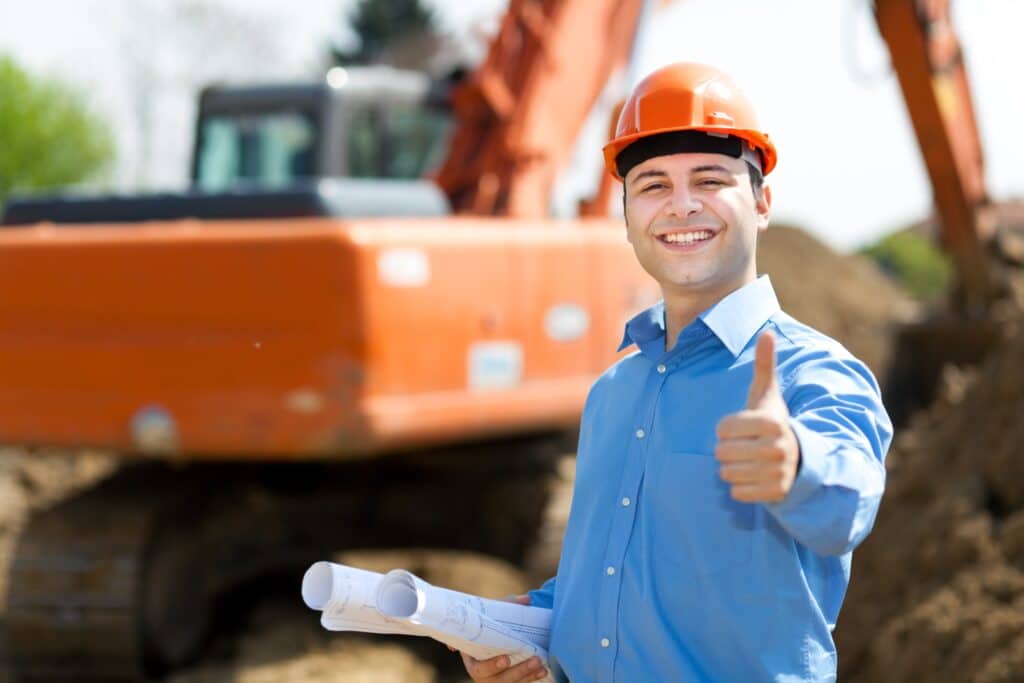 Un ouvrier portant une chemise bleue et une casque de protection, souriant et levant le pouce et tenant dans son autre main un document.