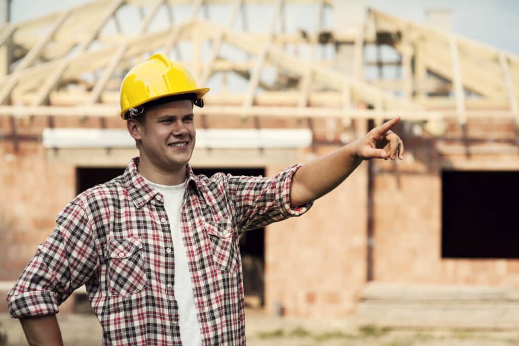 Un ouvrier portant un casque jaune et une chemise à carreaux se tenant devant une maison en cours de construction et pointant un doigt devant lui.