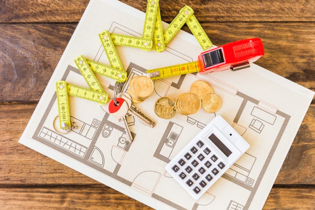 Un plan, un double décamètre, une calculatrice, des clés et des pièces de monnaies, posés sur une surface en bois.