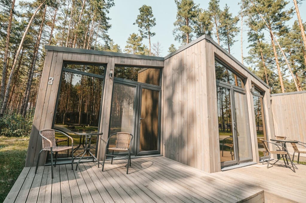 Une maison en bois de forme carré se trouvant dans les bois, avec des baies vitrées et une terrasse en bois.